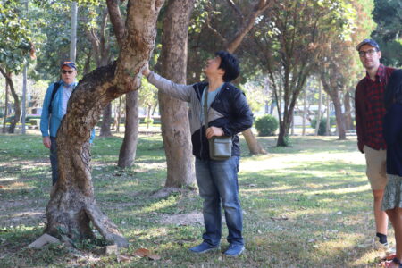 A guide examining a tree during a nature walk at Ang Kaew Reservoir, with participants observing in the background among a serene wooded area.
