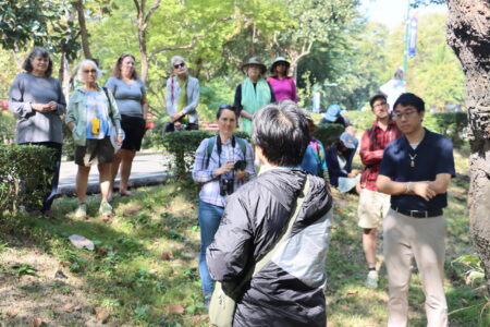 A group of participants attentively listening to a guide during a nature walk at Ang Kaew Reservoir, with the guide facing the group and lush greenery in the background.