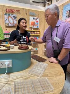 A person is receiving a chocolate-making demonstration, while others observe and learn the process of crafting their own chocolate bars.