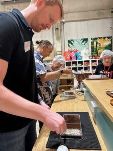 A participant carefully adding ingredients to their chocolate bar mold, surrounded by other participants during the chocolate-making workshop.