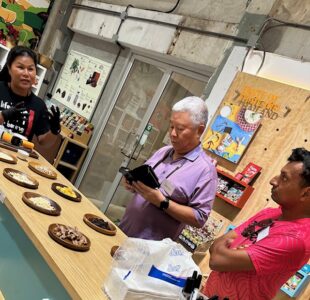 A staff member at Siamaya Chocolate Factory is giving a demonstration to participants, showing various chocolate ingredients laid out on plates, while two participants observe and engage in the discussion.