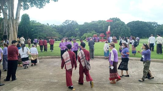 Participants of the Chiang Rai excursion and local community members gather in a large outdoor space, some dressed in traditional attire, others in casual clothing. The scene captures the vibrant atmosphere of the cultural event, with a mix of walking, talking, and preparing for the next activity, symbolizing the lively exchange between visitors and the local community.