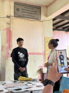 A local community member explains a project as part of the Chiang Rai excursion, with a participant listening attentively. The conversation takes place near a sign about a community development project, illustrating the importance of local initiatives in preserving the environment and culture of the upland communities.