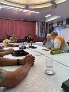 Participants of the Chiang Rai excursion engage in a discussion during a meeting session. One participant speaks into a microphone, while others listen attentively. The image captures the interactive nature of the visit, highlighting the importance of open dialogue and learning from local experts in the region's cultural and social context.