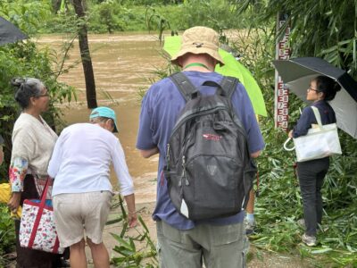 Participants of the Chiang Rai excursion observe the river's water levels during a field visit. Some are gathered near a flood gauge, reflecting the importance of environmental awareness and the impact of natural events on the local communities. The image shows the group, equipped with umbrellas, engaged in learning about local challenges.