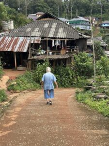 A participant walks through a Karen village during the 4-day, 3-night excursion to Chiang Rai, offering a glimpse into the lifestyle and environment of the upland peoples. The image showcases the rural village setting, with simple wooden homes surrounded by lush greenery, highlighting the close connection between the people and their land.