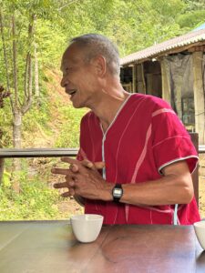 Preecha Siri, a community leader from Huay Hin Lad Nai, shares an engaging moment during the 4-day, 3-night excursion to Chiang Rai. Wearing a red traditional shirt, Preecha enthusiastically interacts with participants, demonstrating the warmth and wisdom of local community leaders who are dedicated to preserving nature and cultural heritage.