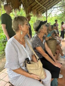 A group of excursion participants sitting under a thatched roof, attentively listening during a presentation at the tea plantation. The scene shows the educational part of the Araksa Tea Garden visit.