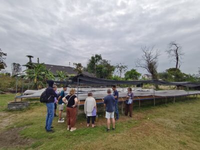The group gathers around raised drying beds under shade netting, learning how beans are dried and prepared before roasting